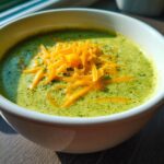 Close-up of a bowl of creamy broccoli cheddar soup, topped with shredded cheddar cheese.