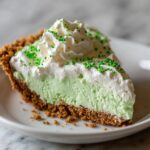 A slice of No Bake Shamrock Shake Pie on a white plate, topped with whipped cream and green sprinkles.