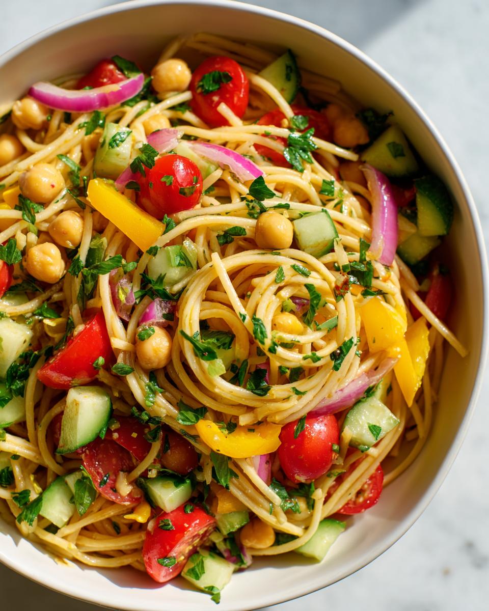 Overhead shot of a delicious pasta salad chickpeas in a white bowl, with fresh vegetables and herbs.