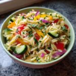 Close-up of a bowl of pasta salad orzo with cucumbers, bell peppers, red onion, and herbs.