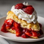 Close-up of a slice of Strawberry Shortcake with fresh strawberries and whipped cream on a white plate.